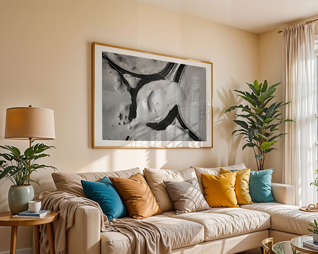 A beige living room features a light sofa with yellow, blue, and gray pillows. Above hangs "Desert Roads Roundabout" by FN Prints, a black and white aerial photo. To the left are a potted plant and lamp, with sunlight streaming in.