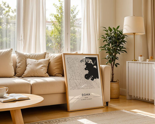 A bright living room features a beige sofa, floor lamp, potted plant, and the FN Prints Doha City Map leaning against the sofa. Sunlight pours in through large windows with sheer curtains.