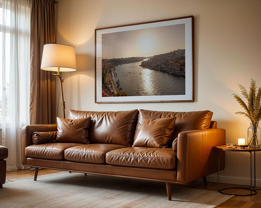 A brown leather sofa with three cushions sits in a living room. Behind it hangs the "Douro River At Sunset" print by FN Prints. To the left is a floor lamp; on the right, a small round table holds a candle and vase.