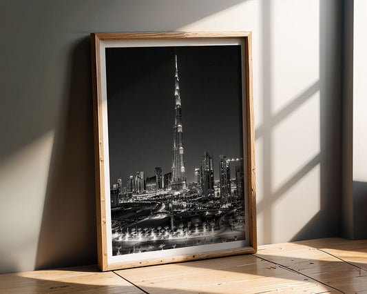 The "Dubai Dreams" framed black and white fine art photograph from FN Prints captures a tall, illuminated skyscraper centered amidst other buildings in the Dubai skyline. The photo is positioned against a wall on a wooden surface, where sunlight creates intriguing shadows over it.