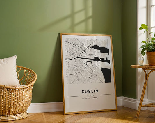 The FN Prints Dublin City Map, framed in black and white with coordinates, leans against a green wall. A wicker chair with a cushion is nearby, and to the right stands a small table with a potted plant. Sunlight highlights the map's minimalist charm through the window shadows.