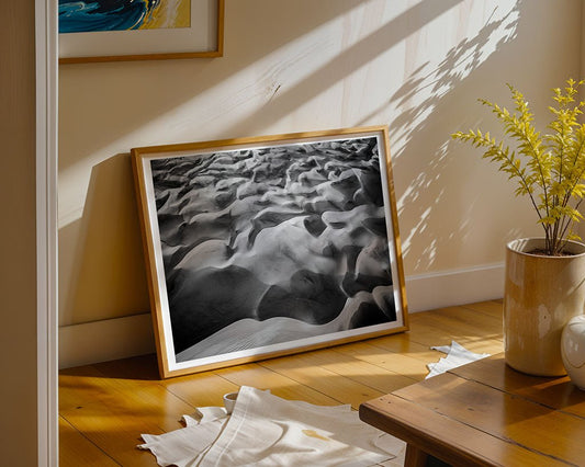 Dune Part IV" by FN Prints, a framed black and white sand dune photo, leans against a beige wall on a wooden floor. Sunlight casts shadows across the scene, with a potted plant and part of a table nearby.