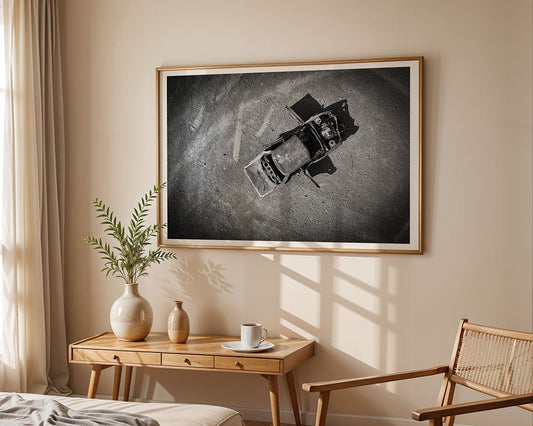 Echoes of Abandonment by FN Prints, a framed black and white artwork of a burnt-out car, hangs on a beige wall above a wooden table set with a vase, mug, and saucer. A wooden chair is nearby as sunlight filters through curtained windows.
