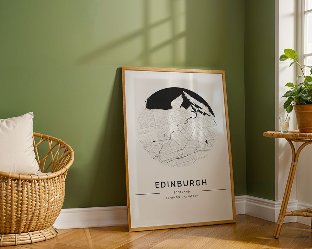 The FN Prints Edinburgh City Map, framed in black and white and printed on museum-quality fine art paper, leans against a green wall beside a wicker chair with a cushion. A small round table with a potted plant adds to the elegance of the wooden floor setting.