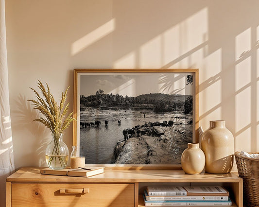“Elephant River” by FN Prints, a framed black-and-white photo of elephants by a river, is displayed on a wooden sideboard with beige and glass vases—bringing serene Sri Lankan wildlife into your home.