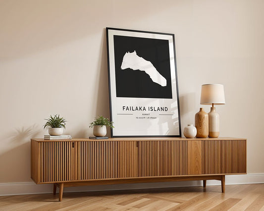 A modern wooden sideboard with vertical grooves stands against a beige wall. On top, there's a framed black and white Failaka Island City Map by FN Prints, two small potted plants, and a table lamp with a wooden base. The floor is light hardwood.