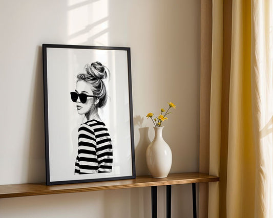 The “French Girl Energy” print by FN Prints features a woman in sunglasses and a striped shirt, styled as black-and-white wall art, bringing Parisian charm to your space when displayed beside décor like vases and flowers.
