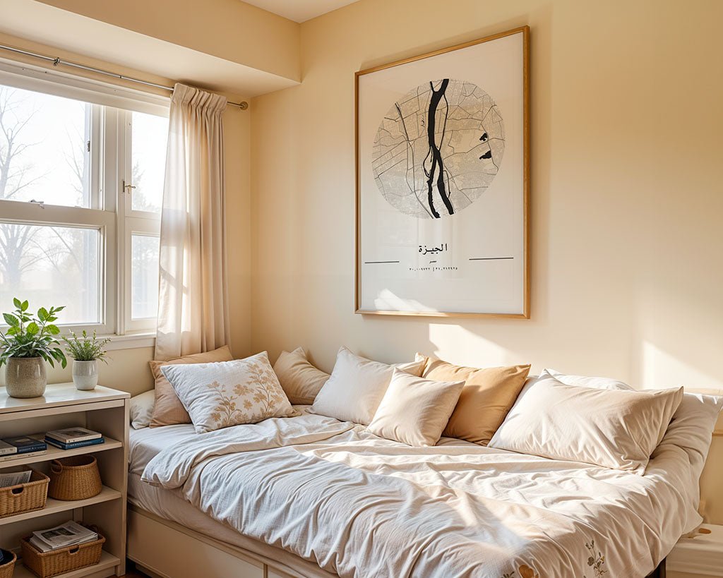 A cozy bedroom corner features a bed with beige and white bedding, numerous pillows, and a large FN Prints Giza City Map on museum-quality fine art paper above. A window with sheer curtains lets in light. Plants and baskets on a small shelf complete the room's warm, inviting atmosphere.