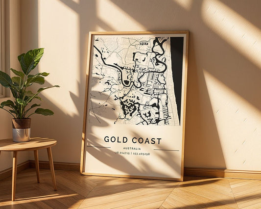 An FN Prints Gold Coast City Map in a minimalist frame leans against a cream wall. Sunlight casts shadows on the floor, where a small green plant sits atop a wooden stool next to the map, perfectly enhancing contemporary interiors.
