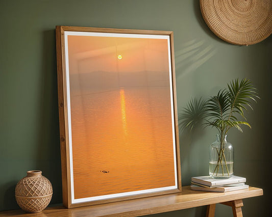 Golden Horizon II" by FN Prints, a stunning Dead Sea sunset photo, leans against a green wall on a wooden table with a leafy plant in a glass vase and a woven basket, as woven wall decor hangs elegantly above.