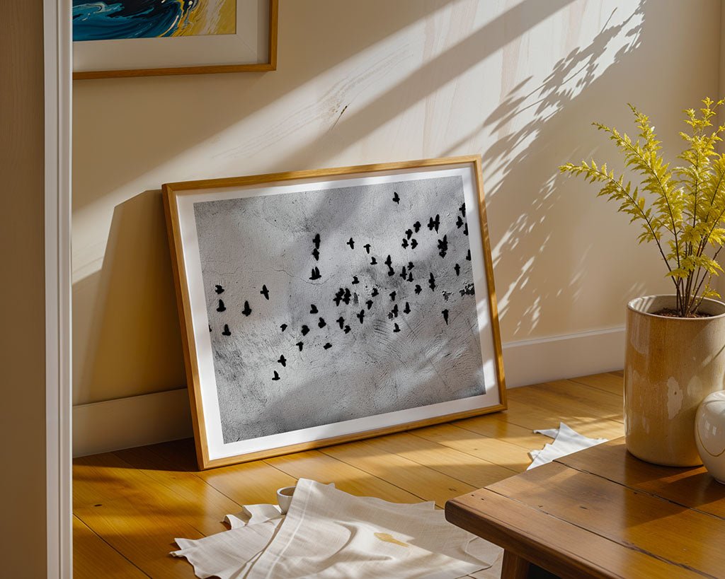 The FN Prints "Graffiti Birds" framed art print against a light wall casts shadows on the wooden floor, complementing the minimalist decor. A small potted plant is on a nearby wooden table, and a white cloth lies on the floor.