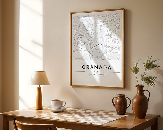 A minimalist room showcases a wooden table against a beige wall. The table holds a lamp, a cup, and two brown vases. A framed FN Prints Granada City Map adorns the wall while sunlight casts artistic shadows across the scene.