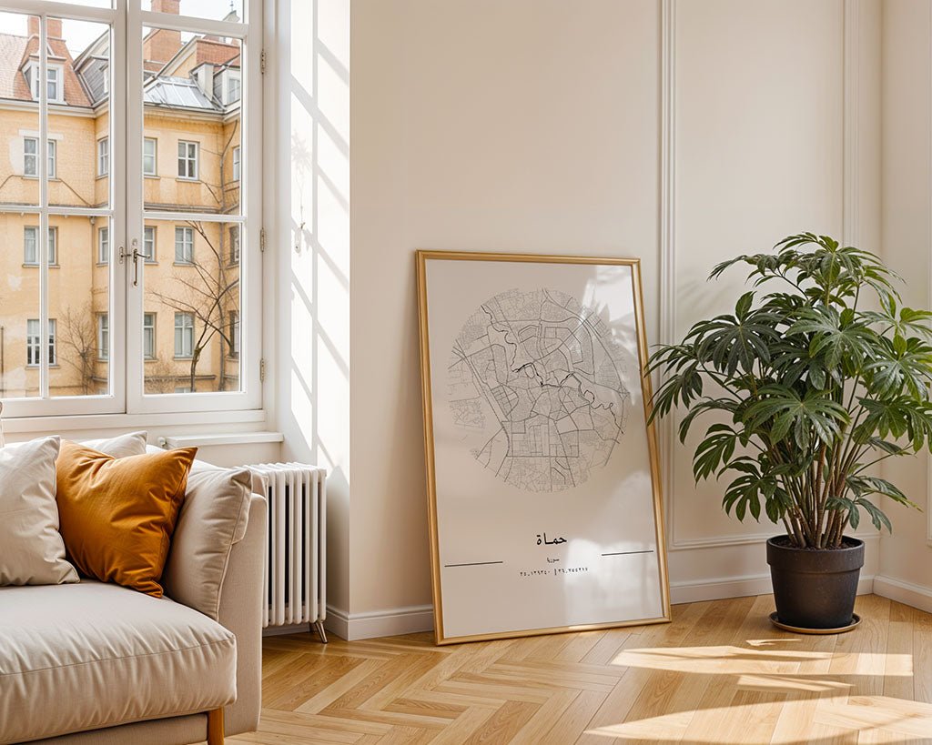 A bright room with a large window reveals neighboring buildings. A minimalist FN Prints Hama City Map leans against the wall while a potted plant sits on the right. On the left, a beige sofa with orange cushions rests, and a herringbone pattern graces the floor.