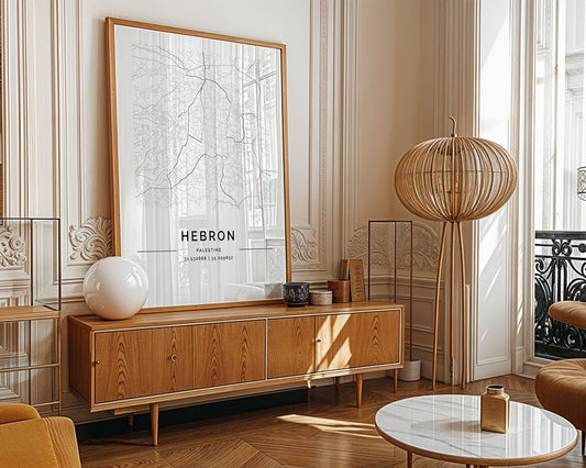 The room exudes minimalist elegance with a wooden console table, decorative items, and a captivating FN Prints Hebron City Map wall art. A unique rattan floor lamp stands beside a round coffee table, while light streams in from the window, casting subtle shadows.