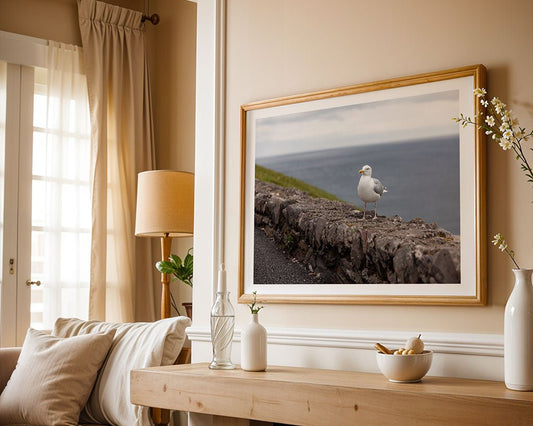 A framed "Herring Gull" photograph by FN Prints, showing a seagull on a stone wall by the sea, hangs on a beige living room wall above a sofa with a lamp and vases arranged on a white shelf nearby.