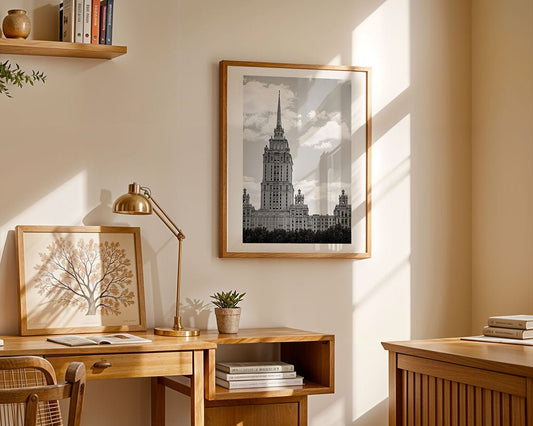 A home office with wooden furniture, a desk lamp, potted plants, books, and framed art—including FN Prints’ “Hotel Ukraina, Moscow.” Sunlight streams through the window, casting shadows on the light-colored walls and furniture.