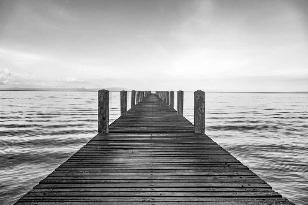 Black and white fine art photography of a long wooden pier stretching into a calm, misty sea under a vast sky in Thailand.