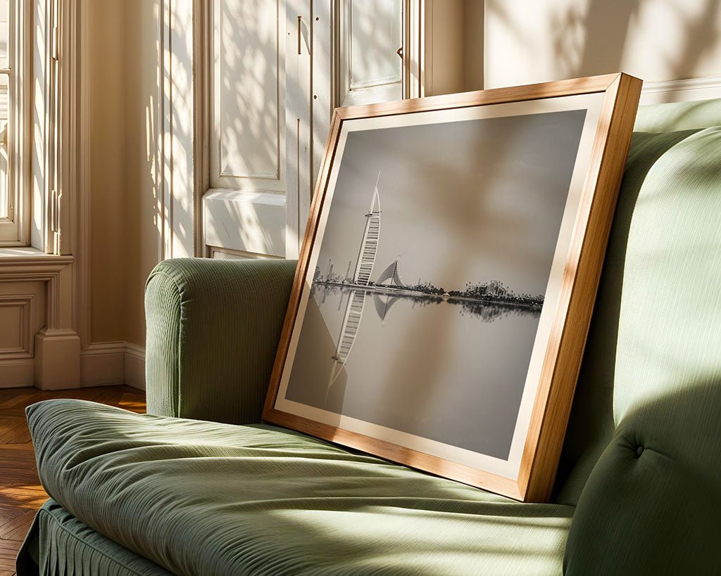 A framed photograph titled "Jumeriah Icons" from FN Prints, showcasing a black and white cityscape with Dubai's iconic sail-shaped skyscraper, reminiscent of the Burj al Arab, is propped on a green sofa. Sunlight streams through nearby windows, creating shadows on the wall and floor.