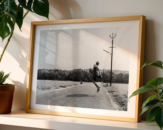 Jumping With Joy" by FN Prints, a framed black and white photo of a joyful woman leaping on a rural road, is displayed on a white shelf with potted green plants as sunlight casts shadows on the wall.