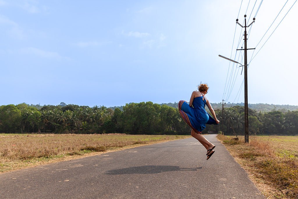 A woman in a blue dress jumping for joy on an empty road lined with tall coconut palm trees in the rural landscape of Goa, India.