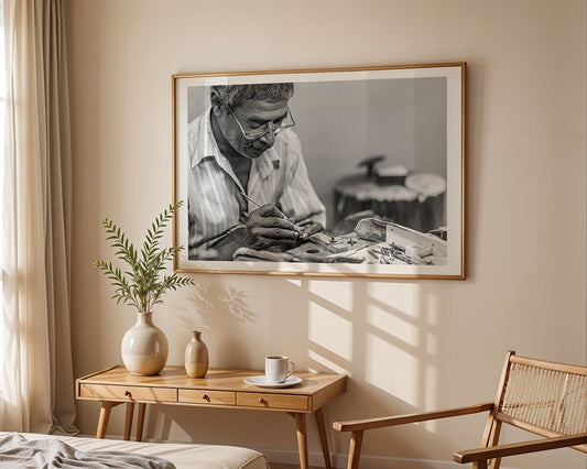 A room with a wooden table and chair creates a serene setting. On the table, there's a vase with green leaves, a cup, and saucer. A framed FN Prints "Kandy Jeweller," featuring a black-and-white photo of a jewellery maker at work, adorns the wall while sunlight casts window shadows.
