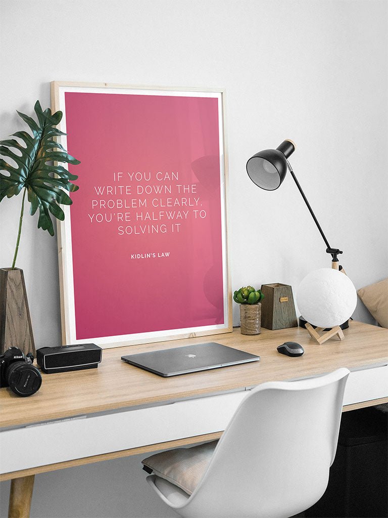 A tidy desk with a white chair, closed laptop, and lamp features FN Prints’ framed pink "Kidlins Law" poster: "If you can write down the problem clearly, you’re halfway to solving it.