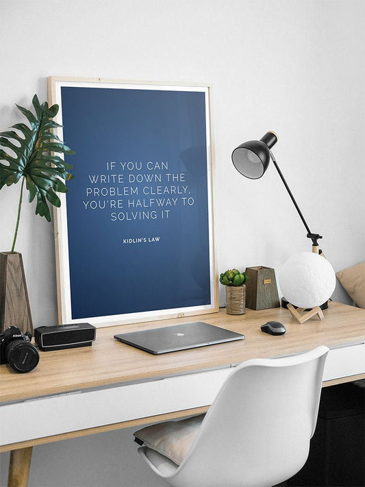 A tidy desk with a white chair, wooden surface, laptop, lamp, small plants, and FN Prints’ Kidlins Law wall art—a motivational quote that reads: "IF YOU CAN WRITE DOWN THE PROBLEM CLEARLY, YOU'RE HALFWAY TO SOLVING IT.