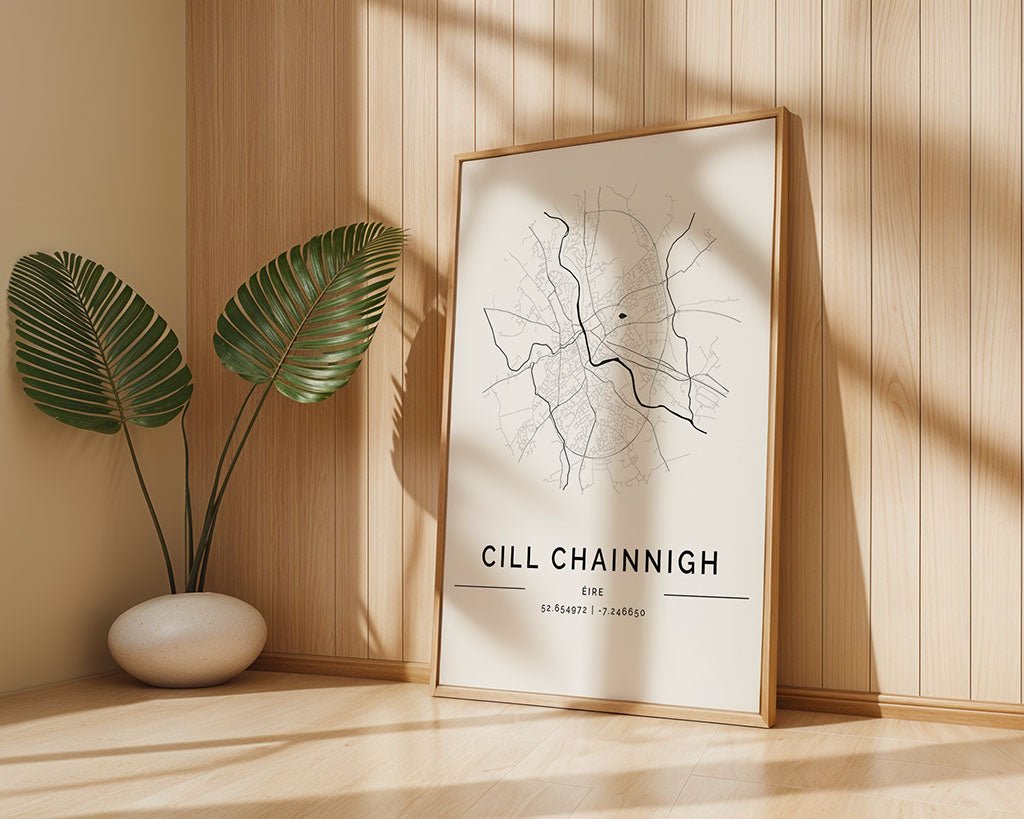 The FN Prints Kilkenny City Map poster, featuring "CILL CHAINNIGH," rests against a light wooden wall. Printed on museum-quality fine art paper, it is paired with two large green leaves in a white vase. Sunlight and shadows enhance its minimalist charm.