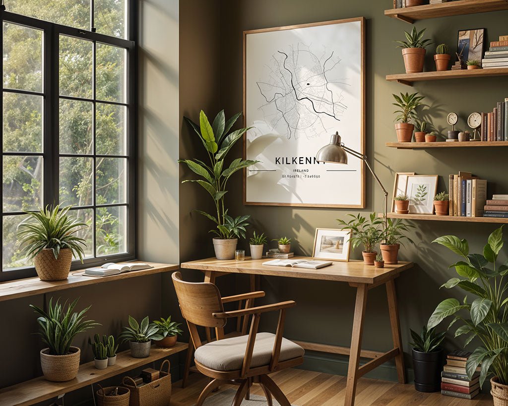 A cozy home office includes a wooden desk and chair, numerous potted plants, and shelves filled with books. A large window on the left brightens a minimalist print of the Kilkenny City Map by FN Prints on museum-quality fine art paper.