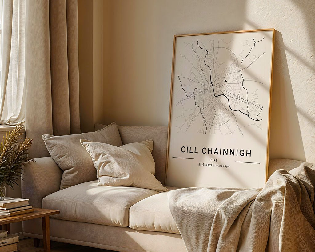A cozy living room showcases a light beige sofa adorned with cushions and a throw. A large framed FN Prints Kilkenny City Map, printed on museum-quality fine art paper, leans against the sofa's back. Sunlight filters through the window, casting warm tones throughout the space.