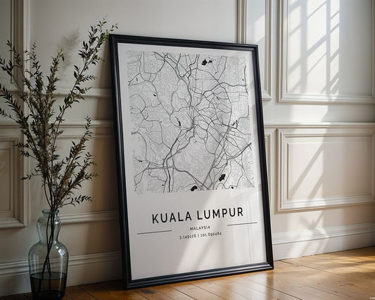 The FN Prints Kuala Lumpur City Map, framed in black and white with coordinates, rests against a white paneled wall, showcasing urban design. A tall glass vase with leafy branches beside it enhances its appeal as wall art on a wooden floor while sunlight casts intriguing shadows.