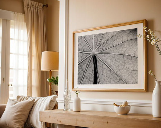 A cozy living room with a beige sofa and wooden table. The wall showcases "Leaf Veins," a large black-and-white macro photograph by FN Prints. A table lamp and decorative vases adorn the table, with cream curtains framing the window on the left.