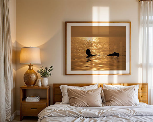 A neatly made bed with beige and white bedding is in a softly lit bedroom. On the wall above the bed hangs “Liquid Gold” by FN Prints, a framed photo of a woman floating in golden water.