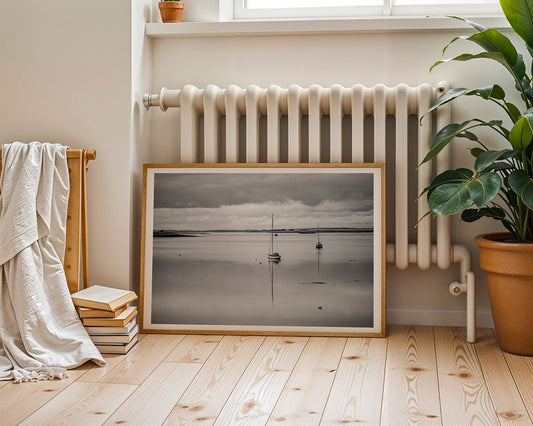 The Lough Derg Black & White art photo by FN Prints features sailboats on calm water, framed and resting against a white radiator on a wooden floor with a potted plant, draped blanket, and stacked books nearby.