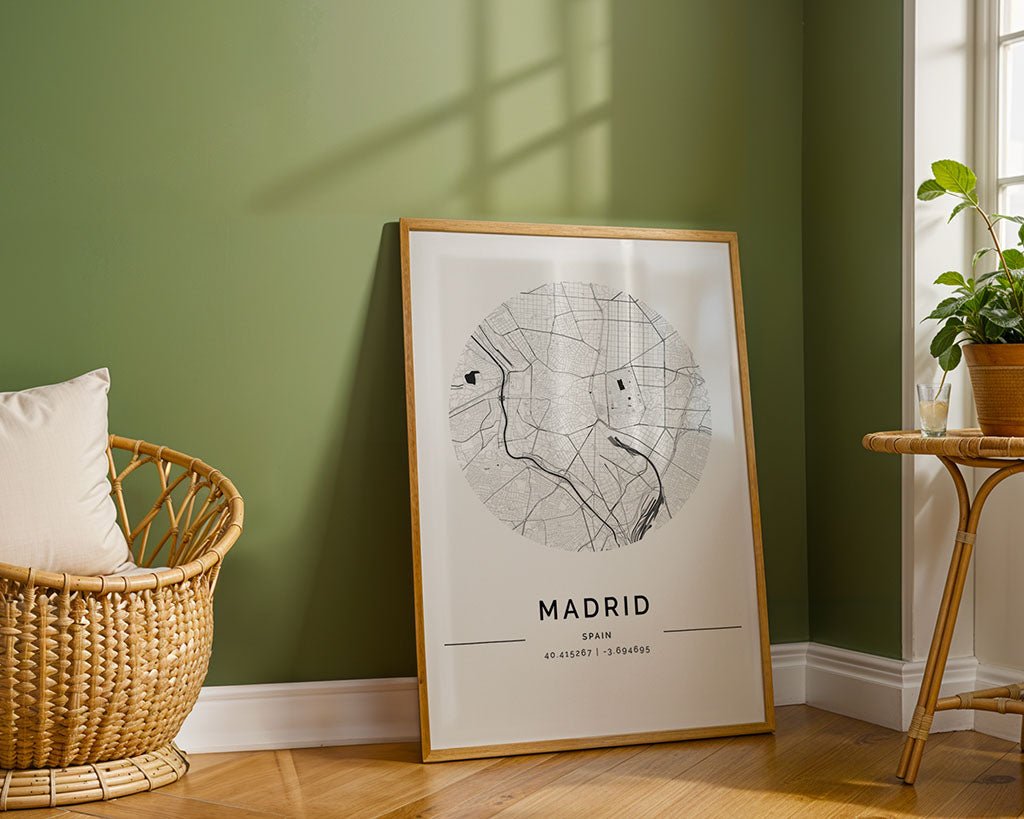The FN Prints Madrid City Map, a framed minimalist city map of Madrid, rests on the floor against a green wall near a wicker chair and a table with a potted plant, illuminated by sunlight from a nearby window.