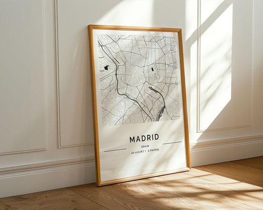 The Madrid City Map by FN Prints, a framed minimalist art piece, leans against a white paneled wall on a wooden floor. Sunlight highlights this elegant Madrid map print, casting gentle shadows across the scene.