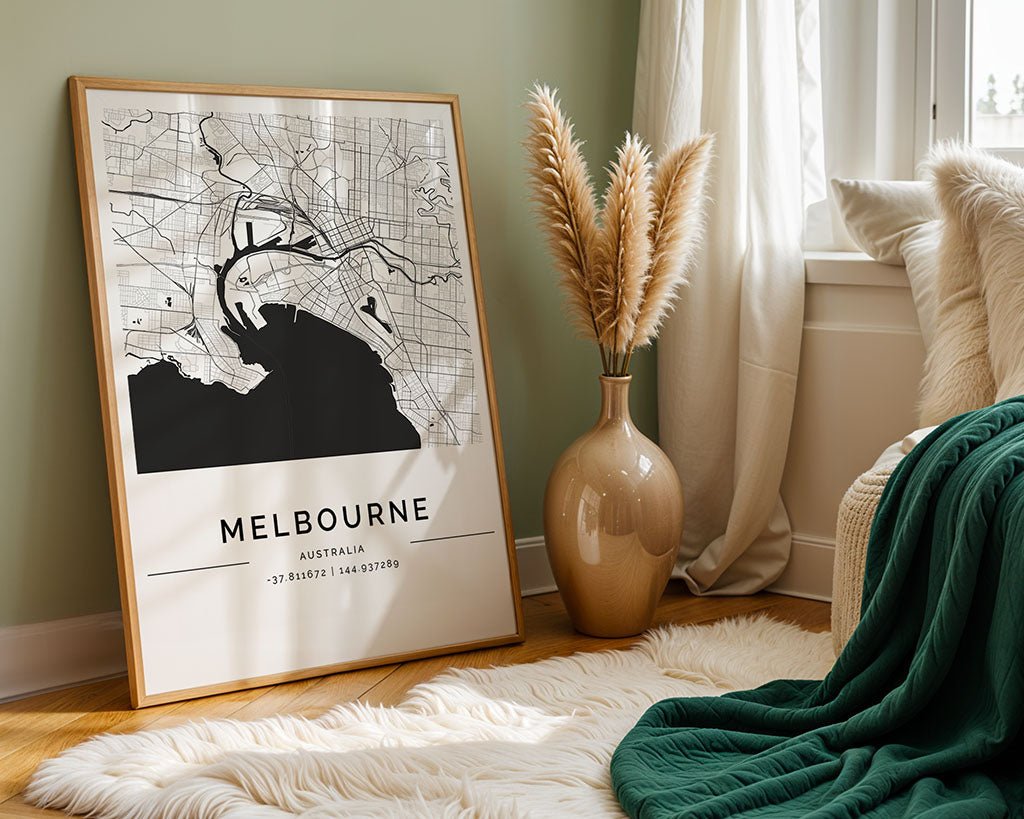 A black and white FN Prints' Melbourne City Map framed poster leans stylishly against a wall with a large beige vase of pampas grass. The space features light green walls, a chair with a green blanket, and a fluffy white rug—perfect for architecture lovers.