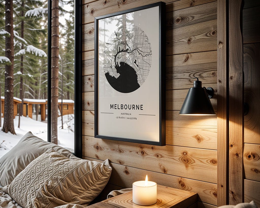 A cozy wooden room showcases FN Prints' minimalist Melbourne City Map, a modern wall lamp, and a small wooden table with a glowing candle. Through the window, snow-covered cabins in the forest are visible. Cream-colored pillows adorn the bed, ideal for architecture lovers.