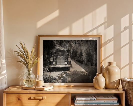 A wooden console displays a FN Prints Mercedes Benz 120 photograph, books, two beige ceramic vases, a glass vase with dried grass, and a notepad. Sunlight highlights this stylish automotive art arrangement.