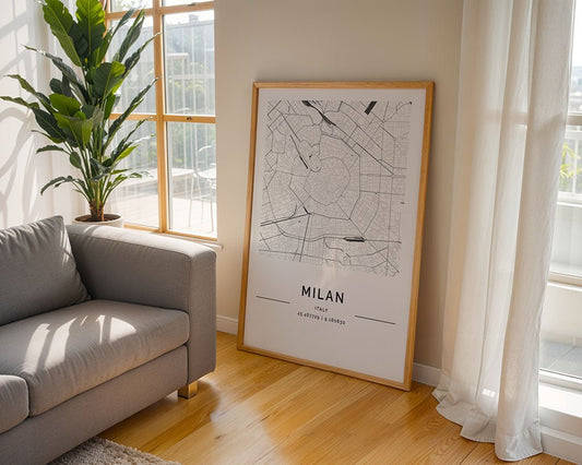 A stylish FN Prints Milan City Map poster, printed on premium paper, is framed against a bright room's wall. It details streets and major areas, complemented by a gray sofa and tall potted plant. Sunlight through large windows enhances the elegant decor.