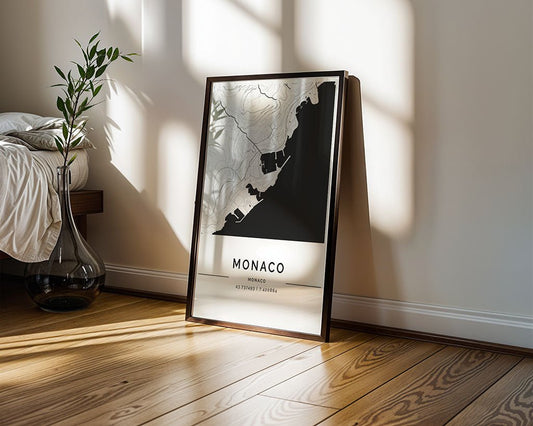 The FN Prints Monaco City Map, displaying black and white geographical contours, leans against a white wall on a wooden floor. Next to it, a large glass vase with green branches is partially visible by the bed, while sunlight enhances its museum-quality paper finish with soft shadows.