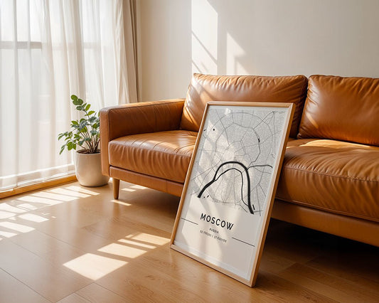 In a sunlit room, a brown leather sofa sits next to a potted plant, with FN Prints' Moscow City Map wall art—an urban design in black and white—resting on the wooden floor. White curtains partially cover the window, allowing soft sunlight to enter.