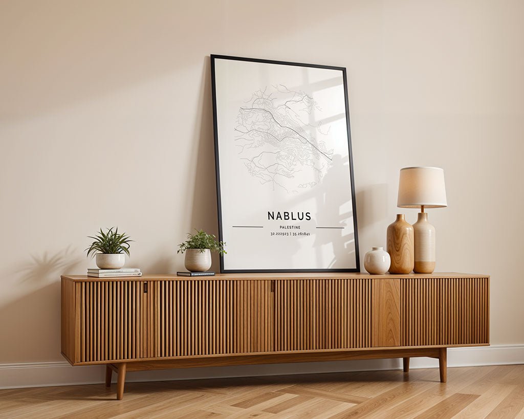 A minimalist wooden sideboard is set against a beige wall, featuring the FN Prints Nablus City Map. It displays two small potted plants, a round vase, and a beige lamp, creating a warm, modern aesthetic.