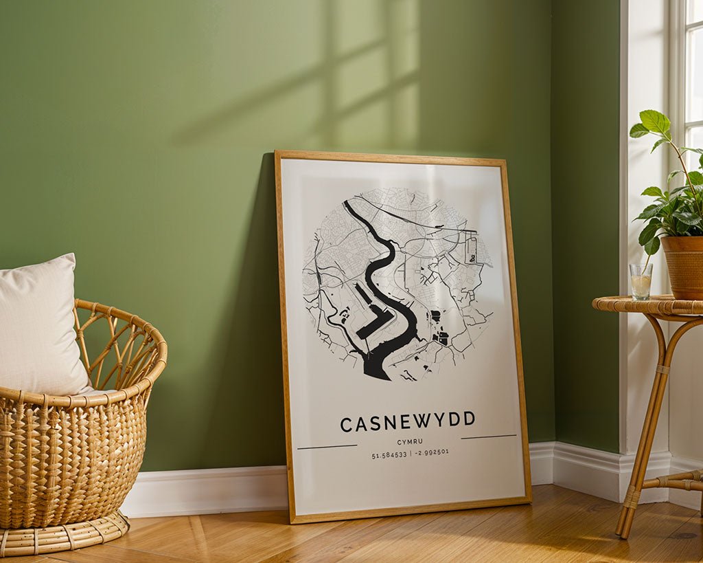 A museum-quality FN Prints Newport City Map leans against a green wall on a wooden floor, showcasing a river and labeled "CASNEWYDD." To the left is a wicker chair with a cushion, and to the right, a small round table enhances its minimalist charm.