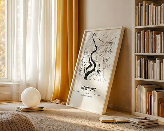 An FN Prints Newport City Map leans against a wall by sheer yellow-curtained windows. A small globe light on stacked books rests on the carpet, while to the right, a bookshelf filled with volumes is illuminated by sunlight, creating a museum-quality fine art ambiance.