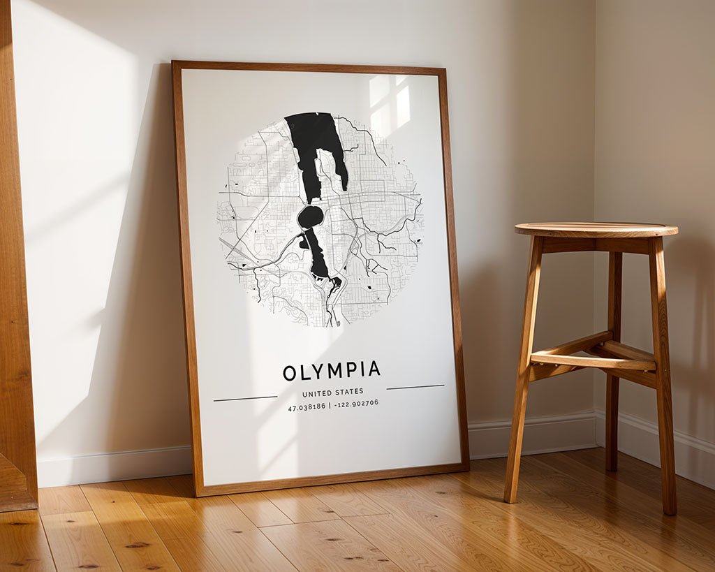 A framed black and white FN Prints Olympia City Map poster is propped against a wall on a wooden floor near a stool. Sunlight enhances the city's allure, while this museum-quality print's detailed geographic coordinates make it a timeless decor piece.