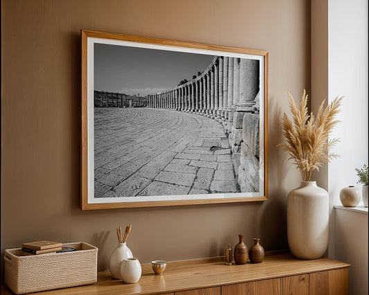 “Oval Plaza Jerash” by FN Prints, a framed black-and-white photo of ancient stone columns and a curved walkway, hangs on a beige wall above a wooden sideboard adorned with vases, a basket, and pampas grass.