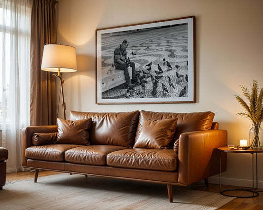 A living room with a tan leather sofa, floor lamp, and side table displays FN Prints' "Pigeon Feeder"—a large framed black and white photo above the sofa capturing the human-animal connection as someone feeds pigeons.