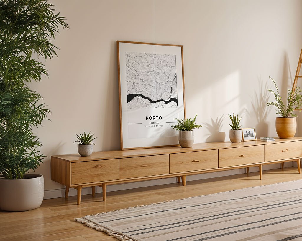 A minimalist living room features a long wooden sideboard with potted plants, a Porto City Map by FN Prints on museum-quality fine art paper leaning against the wall, and to the left, a large plant complements the striped rug as natural light filters in.