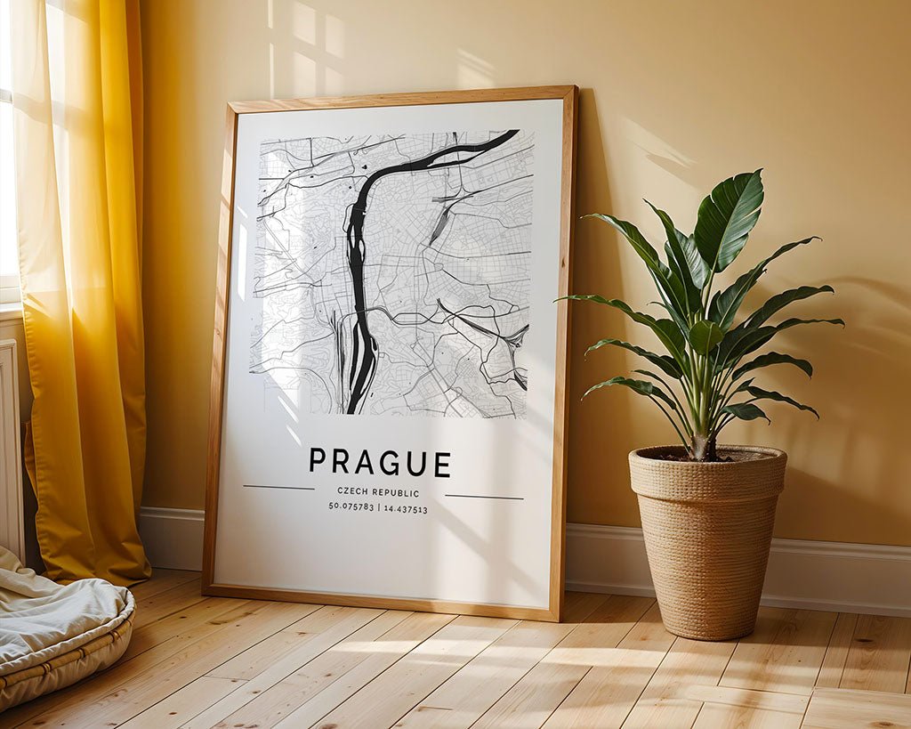 A framed FN Prints Prague City Map leans against a beige wall on a wooden floor, serving as elegant wall art. Beside it, a flourishing potted plant sits under sunlight filtering through a yellow curtain. The map features Prague's coordinates for added charm.